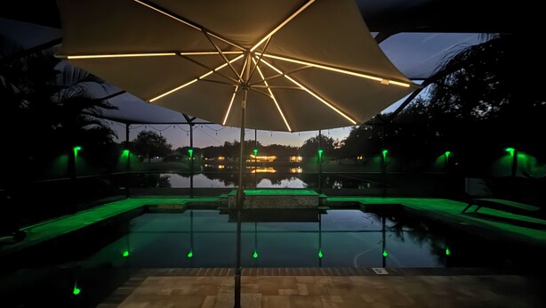 An outdoor swimming pool area with a large umbrella and green ambient lighting. - Relax in my calm and stylish space. Enjoy the relaxing lakeview and heated pool. (Riverview)