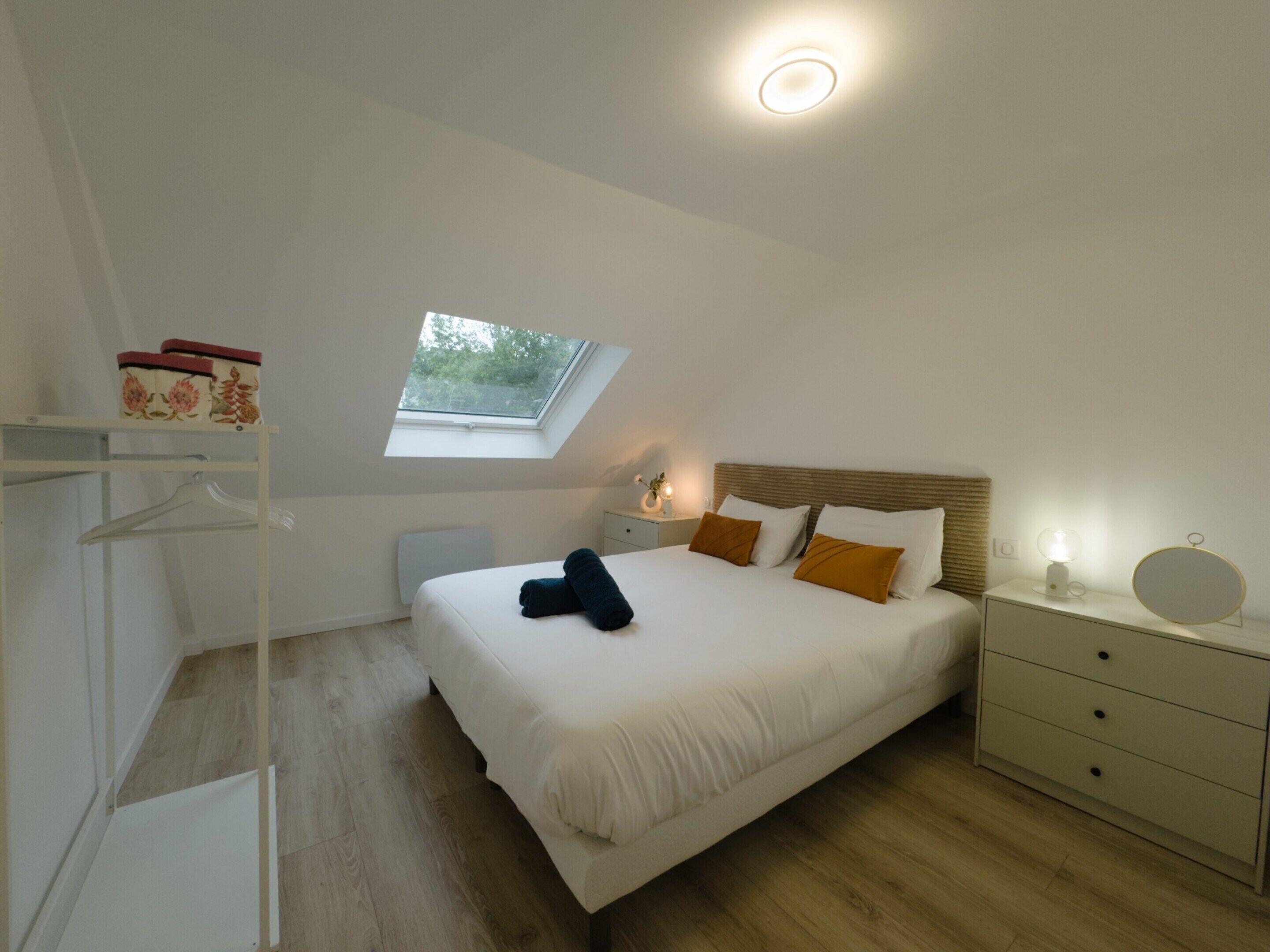 A bedroom with a skylight, a bed with pillows, a bedside table with a lamp, and a wardrobe.