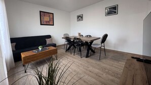 Ein modernes Wohnzimmer mit einem schwarzen Sofa, einem Holztisch und einem Esstisch mit Stühlen. An den Wänden hängen zwei gerahmte Bilder.