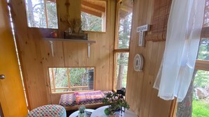 Interior acogedor de una cabaña con paredes de madera, una pequeña zona de estar con cojines, una mesa redonda con una planta y una ventana con vistas a follaje.