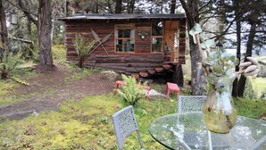 Una cabaña rústica de madera con una mesa y sillas de vidrio al frente, rodeada de árboles y un pequeño jardín.