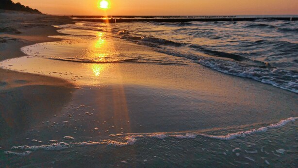 A beach at sunset with the sun low on the horizon, waves gently lapping at the shore, and a pier extending into the water.