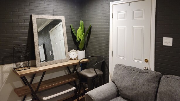 A room with a dark brick wall, a white door, a grey sofa, a wooden table with a mirror, a potted plant, and a small black chair. - Charming 3-bedroom house with AC, WiFi in fabulous Mobile (Mobile)