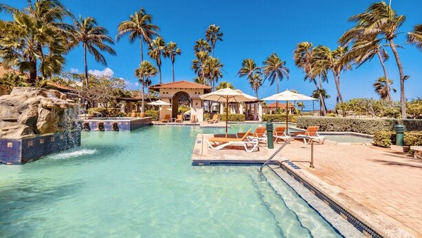 A clear pool with lounge chairs, palm trees, and a building with a tiled roof.