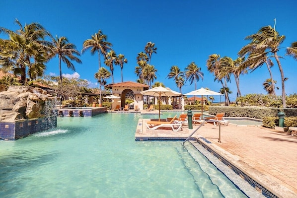 A clear pool with lounge chairs, palm trees, and a building with a tiled roof. - Luxury 3BR with Stunning Views - Anabui 3 Bocobay (Noord)