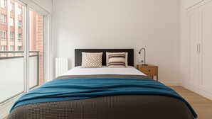 A bedroom with a bed, a nightstand, a lamp, and a view of buildings through a window. - Spacious apartment in the city center (Bilbao)