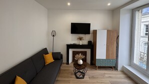 Un salon moderne avec un canapé noir, une petite table basse, une cheminée, un téléviseur fixé au mur et une grande fenêtre.