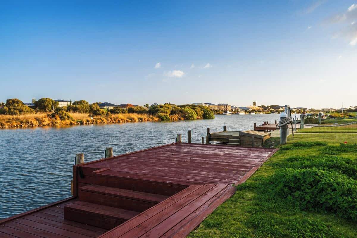 A wooden dock extending into a calm river, surrounded by lush greenery and distant buildings.