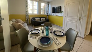 Un área de comedor con una mesa redonda dispuesta para cuatro personas, un sofá, un televisor montado en la pared y una puerta a la derecha.