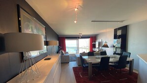 Una sala de estar moderna con comedor, un ventanal con cortina roja y vista al mar.