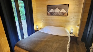 Une pièce chaleureuse, aménagée dans un chalet en bois, avec un lit, une table de chevet, un tableau représentant des montagnes accroché au mur et une fenêtre avec des rideaux.