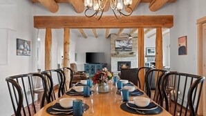 Un'area pranzo con un tavolo di legno apparecchiato per sei persone, circondato da sedie, con un lampadario sopra e un caminetto sullo sfondo.