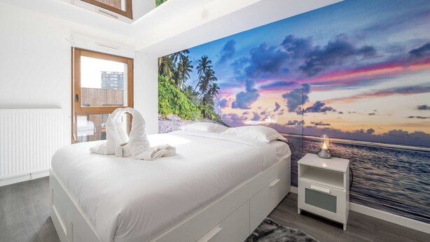 Une chambre avec un grand lit, une table de chevet, une fenêtre et une fresque murale représentant un coucher de soleil sur l’eau.