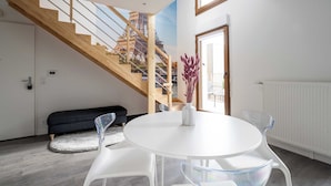 Un salon moderne avec une table ronde blanche, des chaises transparentes, un canapé noir et un escalier en bois menant à l’étage supérieur.
