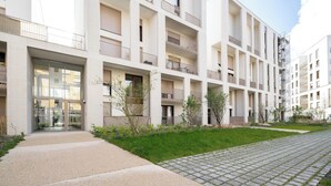 Un immeuble résidentiel moderne à plusieurs étages, doté de balcons, d’une entrée pavée et d’un petit espace verdoyant.