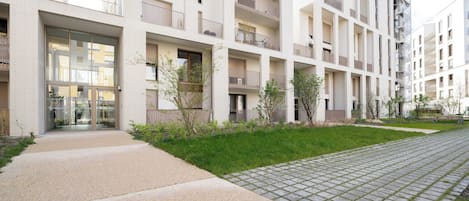 Un moderno edificio residencial de varios pisos, con balcones, entrada pavimentada y un pequeño espacio con césped.