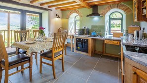Una cocina rústica con vigas de madera, un comedor con mesa para cuatro, y vistas al campo a través de amplias ventanas.
