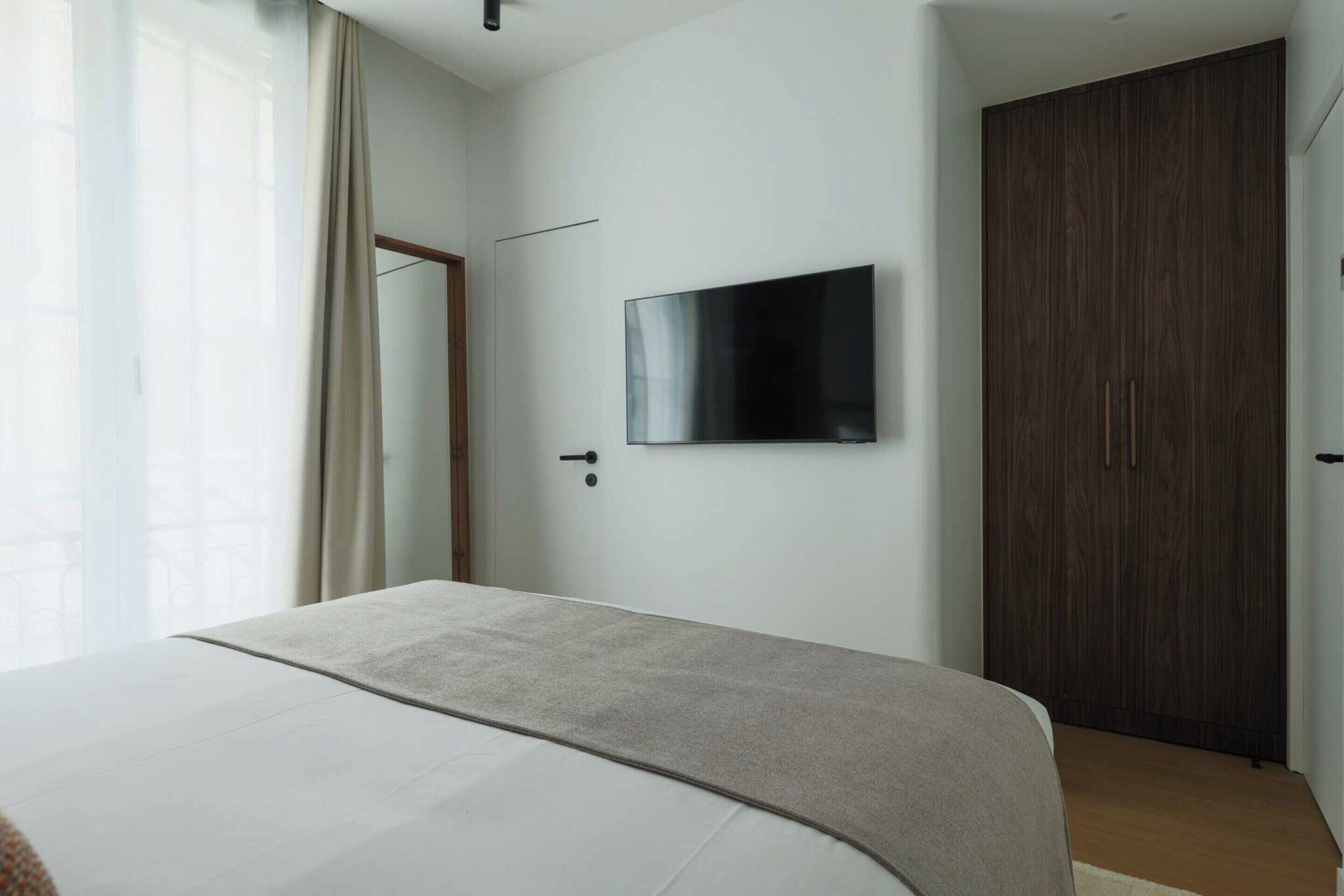 A modern bedroom with a bed, a TV mounted on the wall, and a wooden wardrobe.