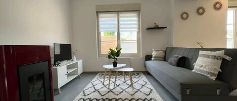 A modern living room with a grey sofa, a TV on a white stand, a patterned rug, and a window with blinds.