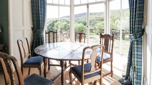 A dining area with a round wooden table and six chairs, large windows with curtains, and a view of greenery outside. - Camisky Lodge (Fort William)