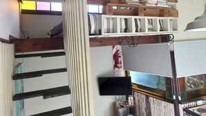 Una habitación con techo inclinado, una escalera de madera, un televisor montado y una pequeña mesa con un extintor.