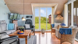 Ein geräumiger Wohnbereich mit Holzboden, einer großen Schiebeglastür, die zu einem Garten führt, einem an der Wand montierten Fernseher und einer gemütlichen Sitzecke mit Sofa und Sesseln.