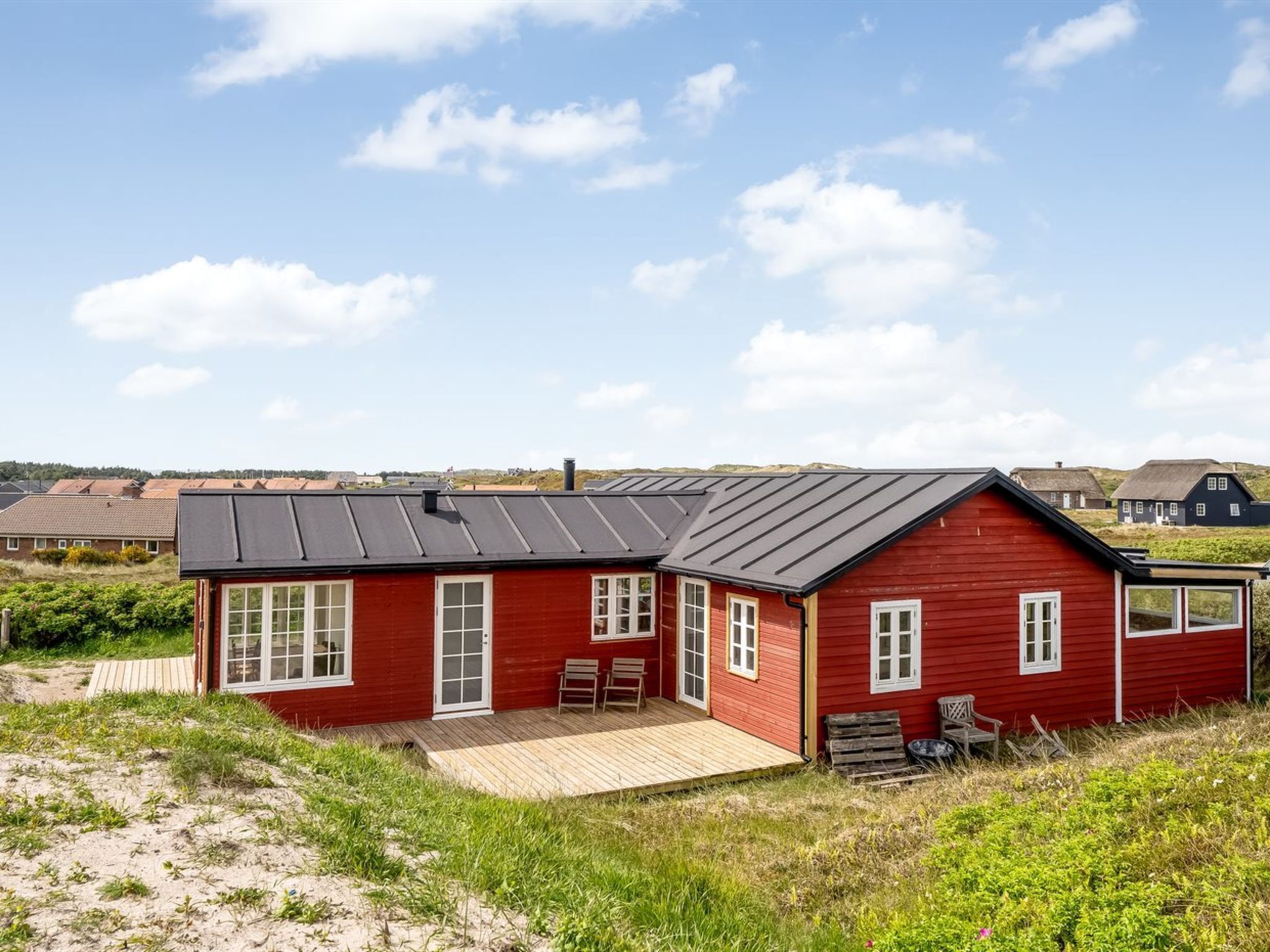 Een rode houten woning met een zwart dak, een houten terras en een klein portaaltje met twee stoelen.
