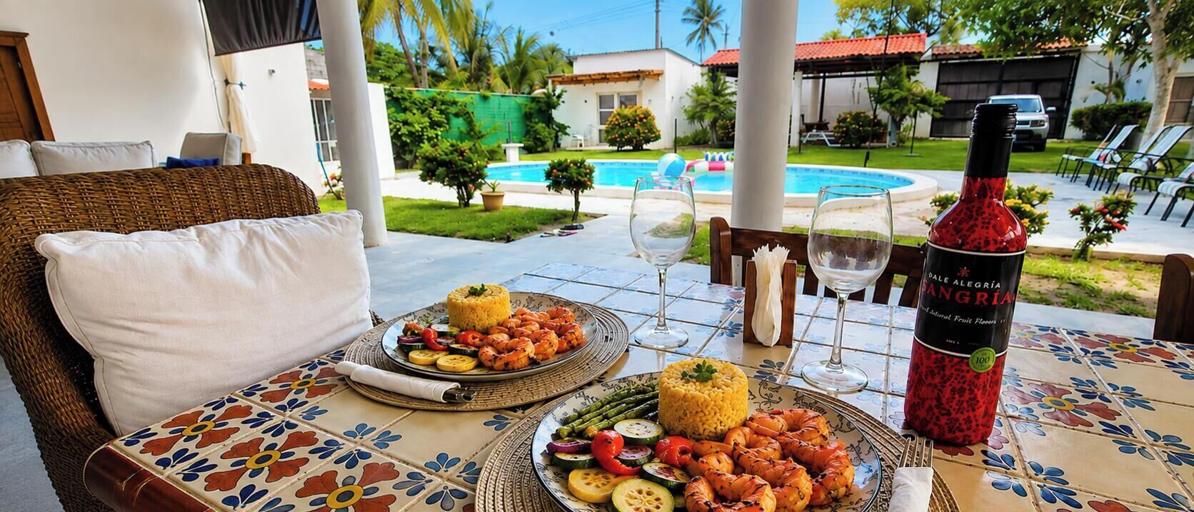 Una mesa de comedor con platos de comida, una botella de vino y vistas a una piscina y zonas verdes.