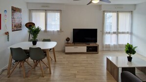 A modern living room with a dining table, chairs, a flat-screen TV, a ceiling fan, and two windows with sheer curtains.