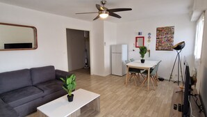 A modern living room with a grey sofa, a wooden coffee table, a dining table with chairs, a ceiling fan, and a wall-mounted TV.