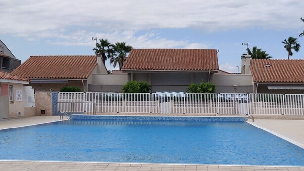 A swimming pool with a tiled deck, surrounded by buildings with red-tiled roofs and a white fence.