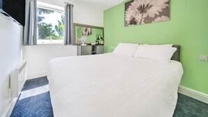 A neatly made bed in a room with a green wall, a window with curtains, a mirror, and a wall-mounted fan. - GO2 Gloucester Hotel by OYO | Standard Double (Gloucester)