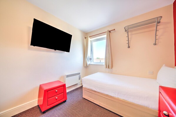 Una habitación pequeña y limpia con una cama, un aparador rojo, un televisor montado en la pared y una ventana con cortinas.