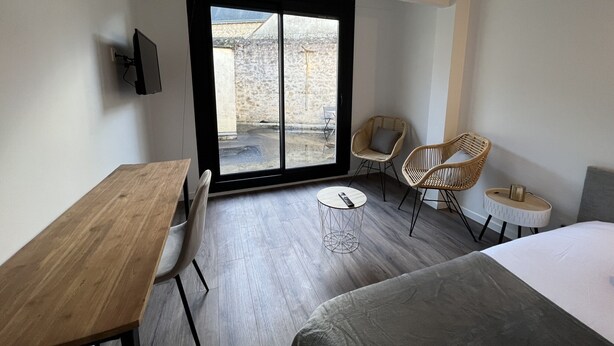 Une chambre d’hôtel moderne, dotée d’un bureau en bois, de deux fauteuils en osier, d’un lit et d’une grande porte-fenêtre coulissante donnant sur un espace extérieur.