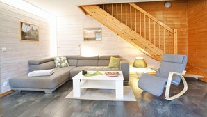 A modern living room with a grey sofa, a white coffee table, and a grey rocking chair. A wooden staircase is visible in the background. - Doppelhaushalfte mit Kamin in Hasselfelde (Oberharz am Brocken)