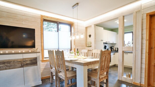 A dining area with a wooden table and chairs, a TV mounted on the wall, and a kitchen in the background. - Doppelhaushalfte mit Kamin in Hasselfelde (Oberharz am Brocken)