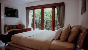 A bedroom with a large bed, a TV mounted on the wall, a sofa with cushions, and a sliding glass door leading to a garden. - Genta Villa Ubud (Ubud)