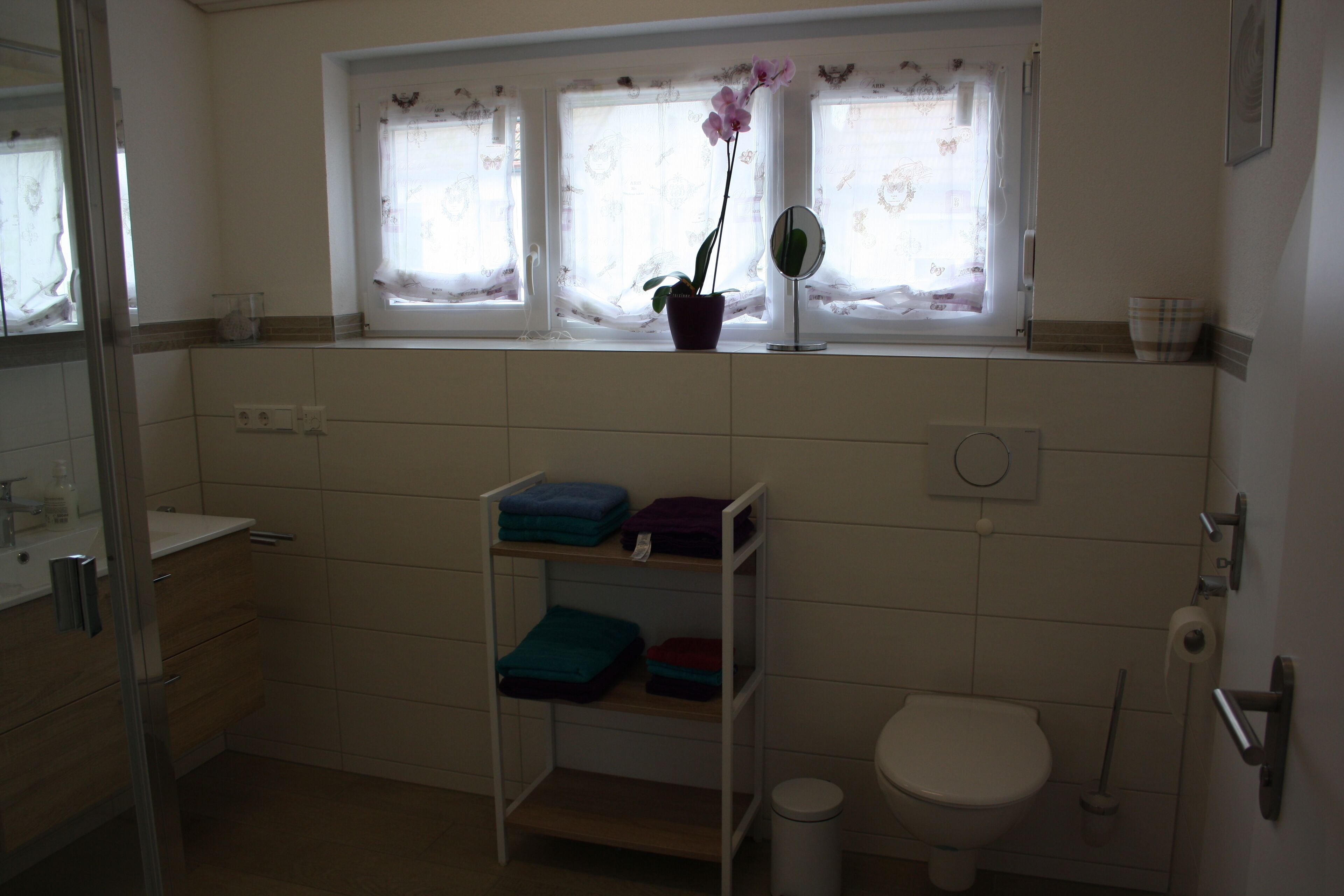 Un baño con inodoro, lavamanos, espejo, estante con toallas dobladas y ventana con cortinas.