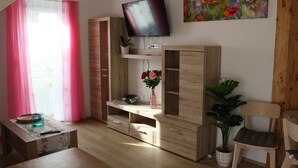 Una sala de estar con un televisor montado en la pared, un mueble de madera para entretenimiento, una silla, una mesa con un jarrón de flores y una ventana con cortinas rosas.