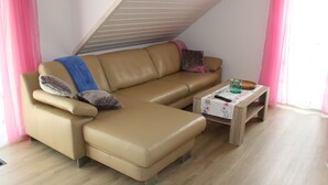 Un sofá de cuero color bronce con una mesa de centro de madera y una mesita auxiliar, techo inclinado y cortinas rosadas.