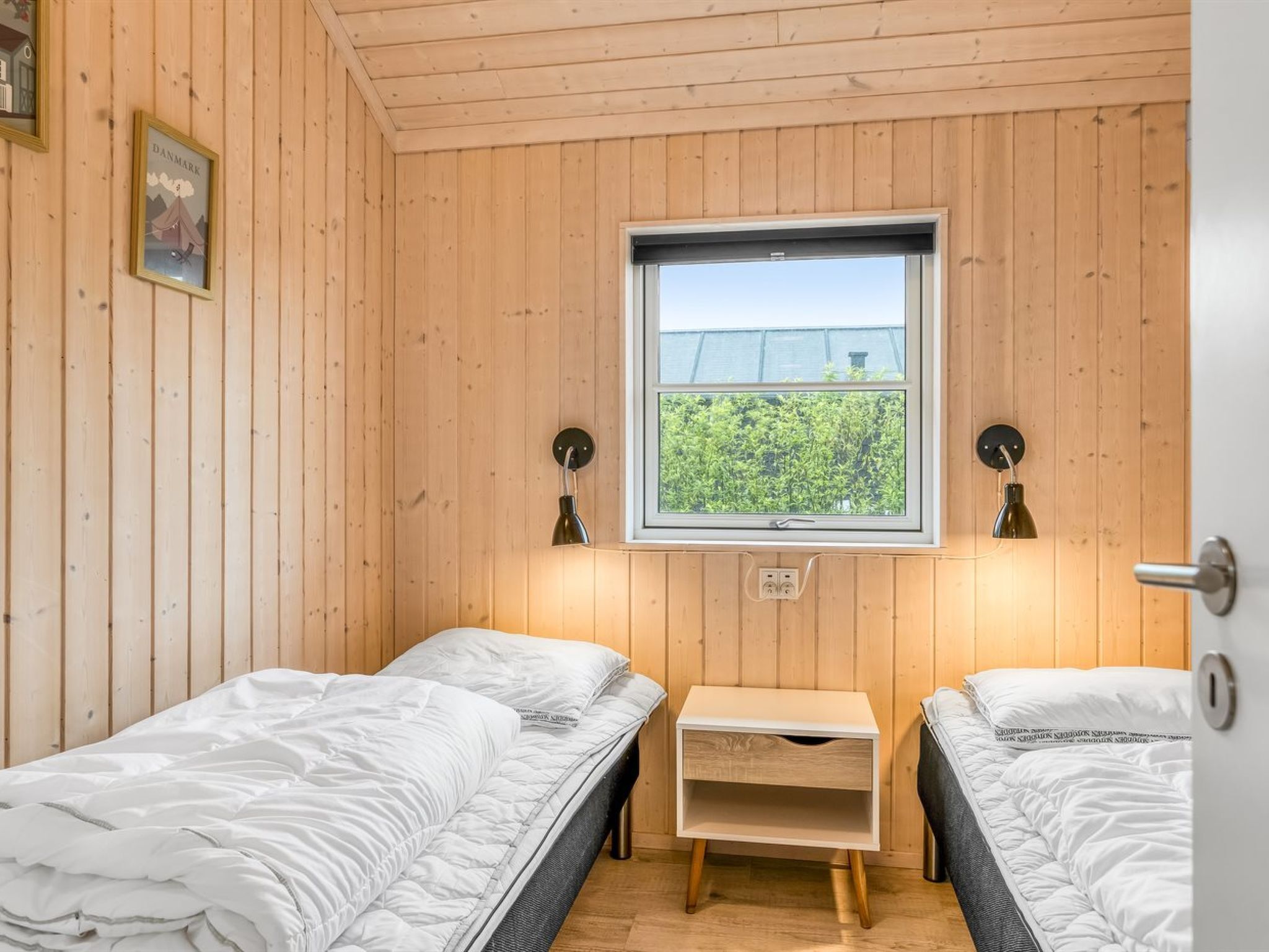 Een knus kamertje met houten wanden en een raam. Er staan twee bedden met wit beddengoed, een klein houten nachtkastje en twee wandlampen.