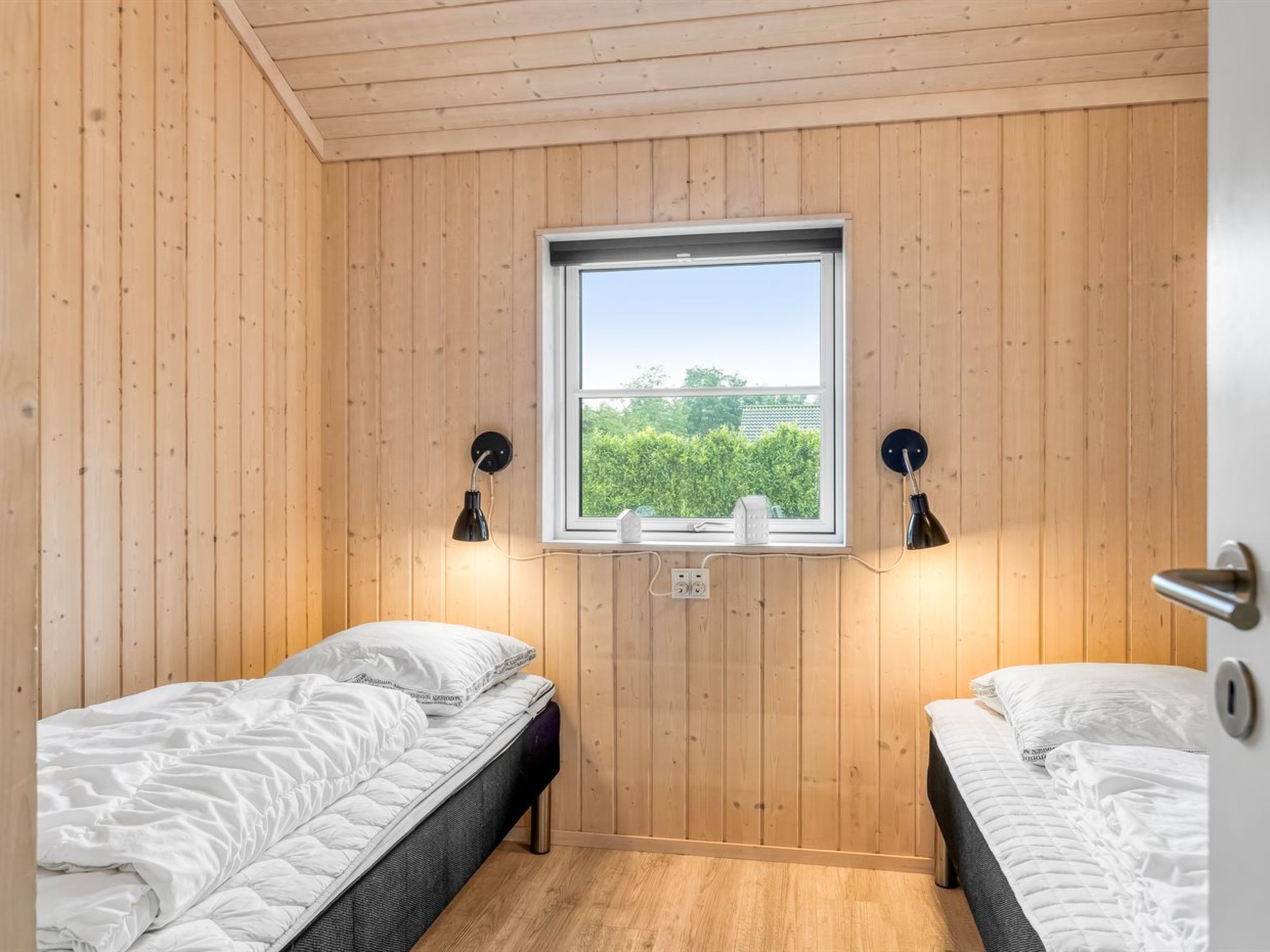 Une petite pièce agréable et chaleureuse, avec deux lits, des murs en bois, une fenêtre donnant sur la verdure et des lampes fixées au mur.