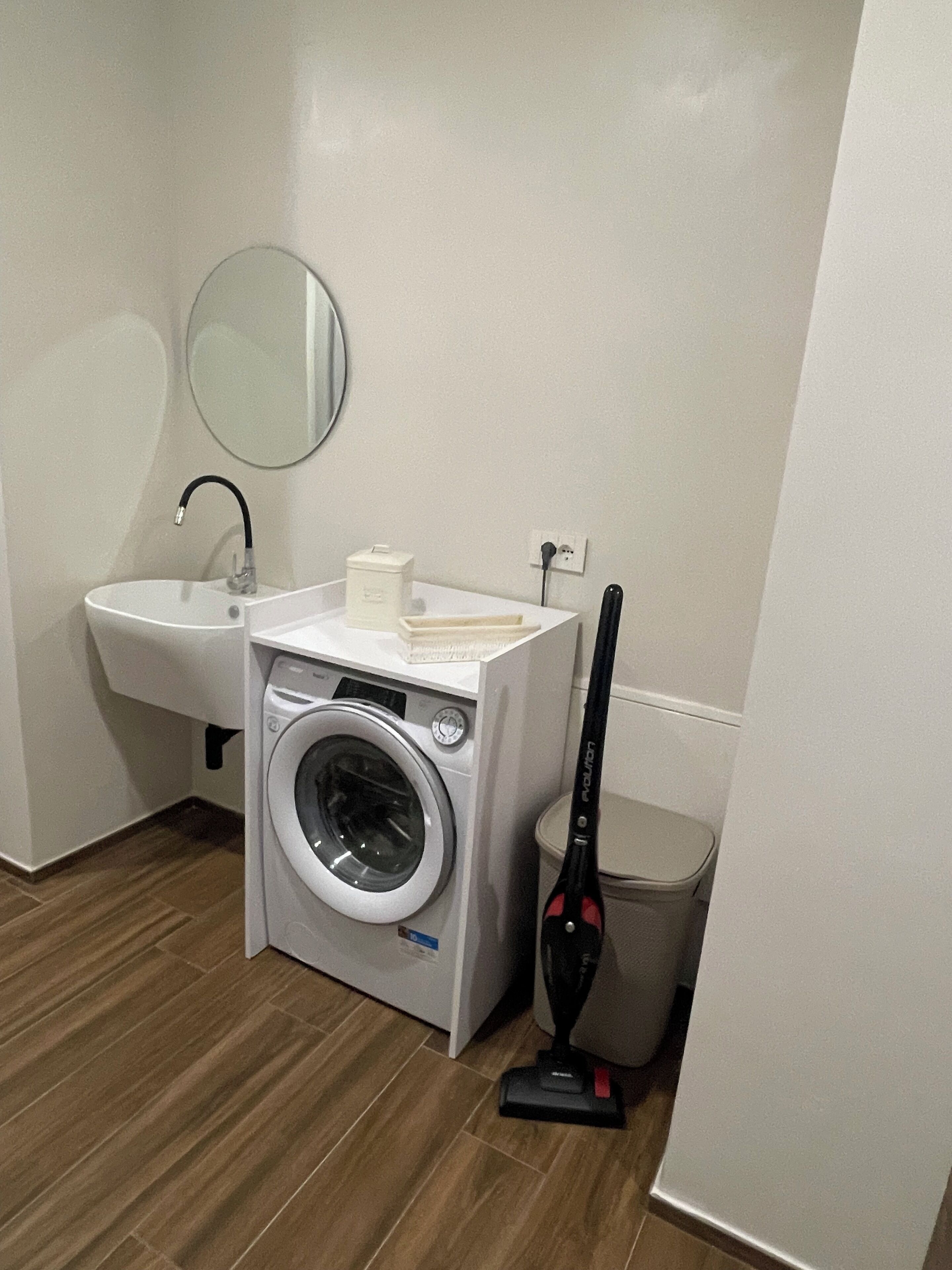 A compact laundry room with a washing machine, a sink, a mirror, and a vacuum cleaner.