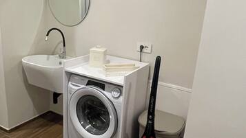 A compact laundry room with a washing machine, a sink, a mirror, and a vacuum cleaner.