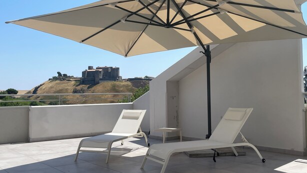 Une terrasse sur le toit, avec deux chaises longues blanches et un grand parasol beige offrant de l’ombre.