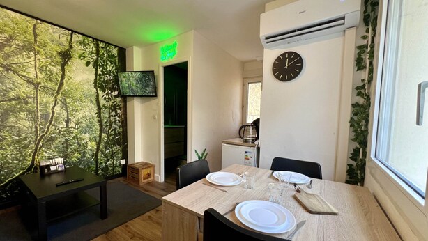 A dining area with a wooden table set for two, a wall-mounted clock, and a television on the wall. - Quiet & Stylish Home With Garden (Le Mans)