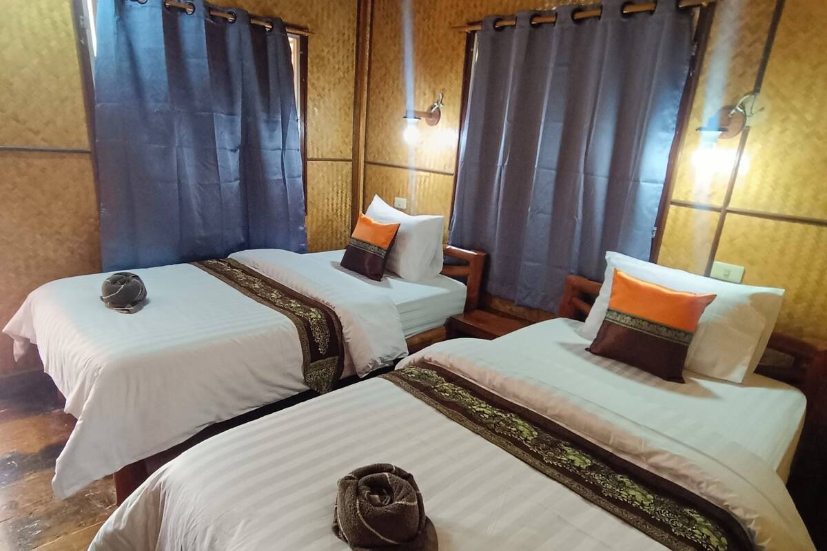 A hotel room with three single beds, each with a neatly folded towel and decorative pillows. The room has wooden walls and a tiled floor.