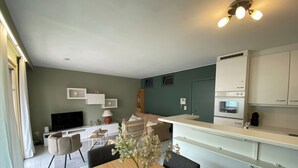 A modern living room with a TV, sofa, and dining table. There is a kitchen with white cabinets and a built-in oven. - Apartment in Nieuwpoort Near the Beach (Nieuwpoort)