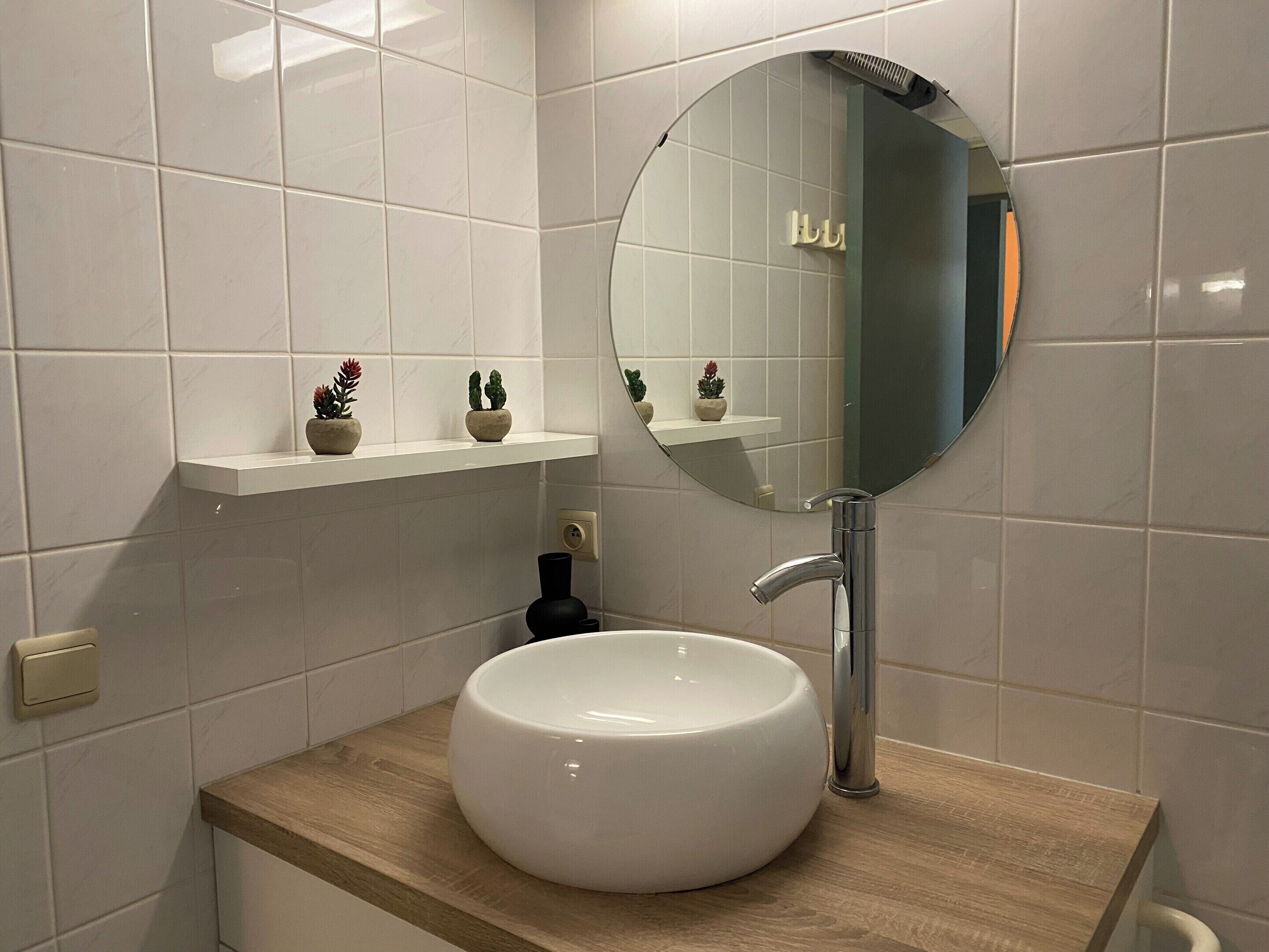 Ein Badezimmer mit einem runden Spiegel, einem weißen Waschbecken und einer Holzauslage. Über dem Waschbecken befinden sich kleine Topfpflanzen auf einem Regal.