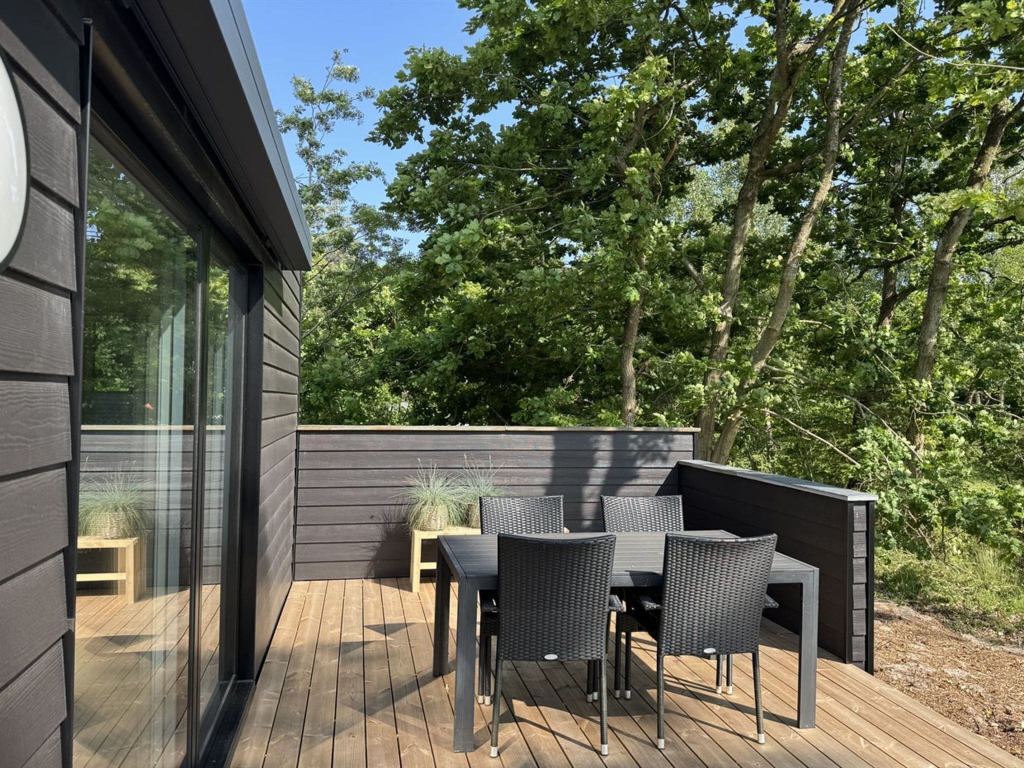 Une terrasse extérieure moderne, dotée d’une plateforme de couleur sombre, d’une porte vitrée donnant sur une pièce et d’un espace détente avec une table et des chaises.